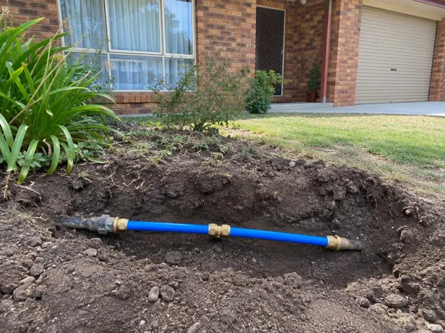 Exposed blue water pipe with brass fittings in a dug trench beside a brick house in Bennetts Green